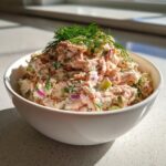 A close-up of creamy salmon salad mixed with red onion and celery, topped with fresh dill, served in a white bowl.