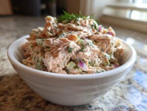 Close-up of creamy salmon salad with celery and red onion, topped with fresh dill, served in a white bowl.