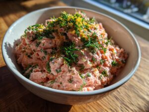 A bowl of creamy, pink salmon dip topped generously with fresh dill and bright yellow lemon zest.