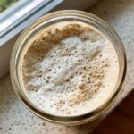 Overhead view of an active sourdough starter in a glass jar, showing a thick layer of foam and many small bubbles.