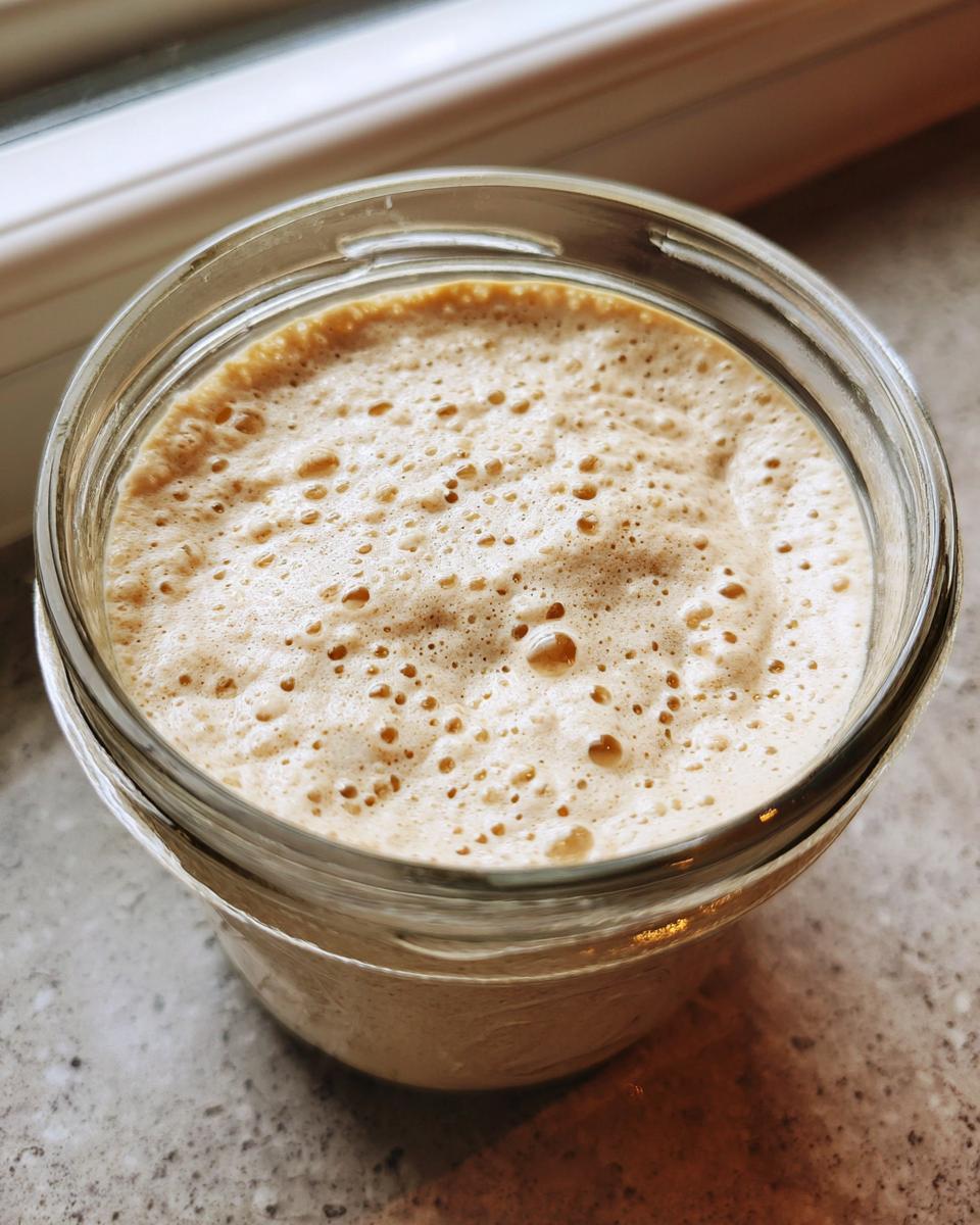 Close-up of an active sourdough starter in a glass jar, showing a thick, bubbly, and foamy top layer.