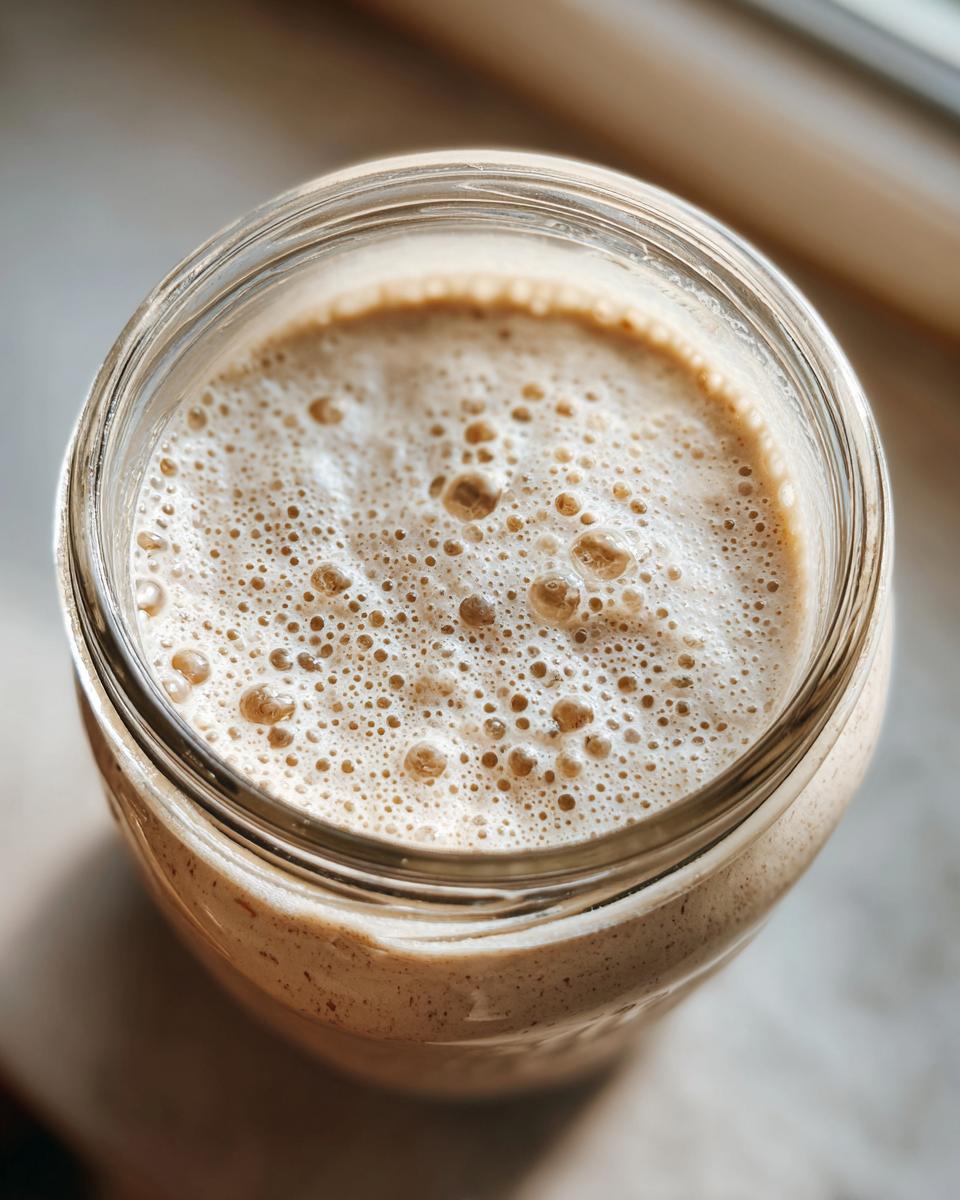 Overhead close-up of an active sourdough starter in a glass jar, showing a thick, foamy top layer full of bubbles.