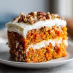 Close-up of a moist slice of carrot cake recipe, layered with cream cheese frosting and topped with pecans.