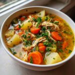 Close-up of a white bowl filled with rich chicken vegetable soup, featuring shredded chicken, carrots, potatoes, and fresh parsley.
