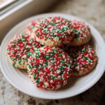 A stack of round christmas sprinkle sugar cookies covered in red, green, and white nonpareils on a white plate.