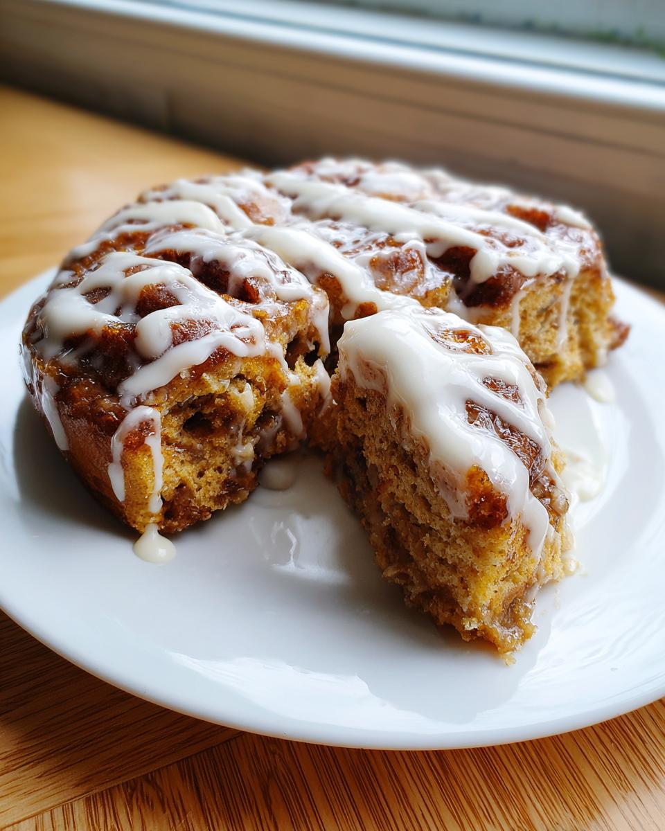 A portion of warm cinnamon roll casserole drizzled heavily with white icing on a white plate.