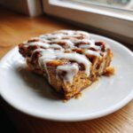 A close-up of a serving of Cinnamon roll casserole drizzled generously with white icing on a white plate.