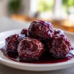 A close-up of several glistening Grape Jelly Meatballs coated in a rich, dark purple sauce on a white plate.