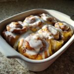 Close-up of soft, golden cinnamon buns baked together in a white dish, generously topped with white icing and cinnamon dust.