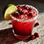 Close-up of a bright red cranberry pomegranate margarita in a salt-rimmed glass, topped with fresh cranberries and ice, garnished with a lime wedge.