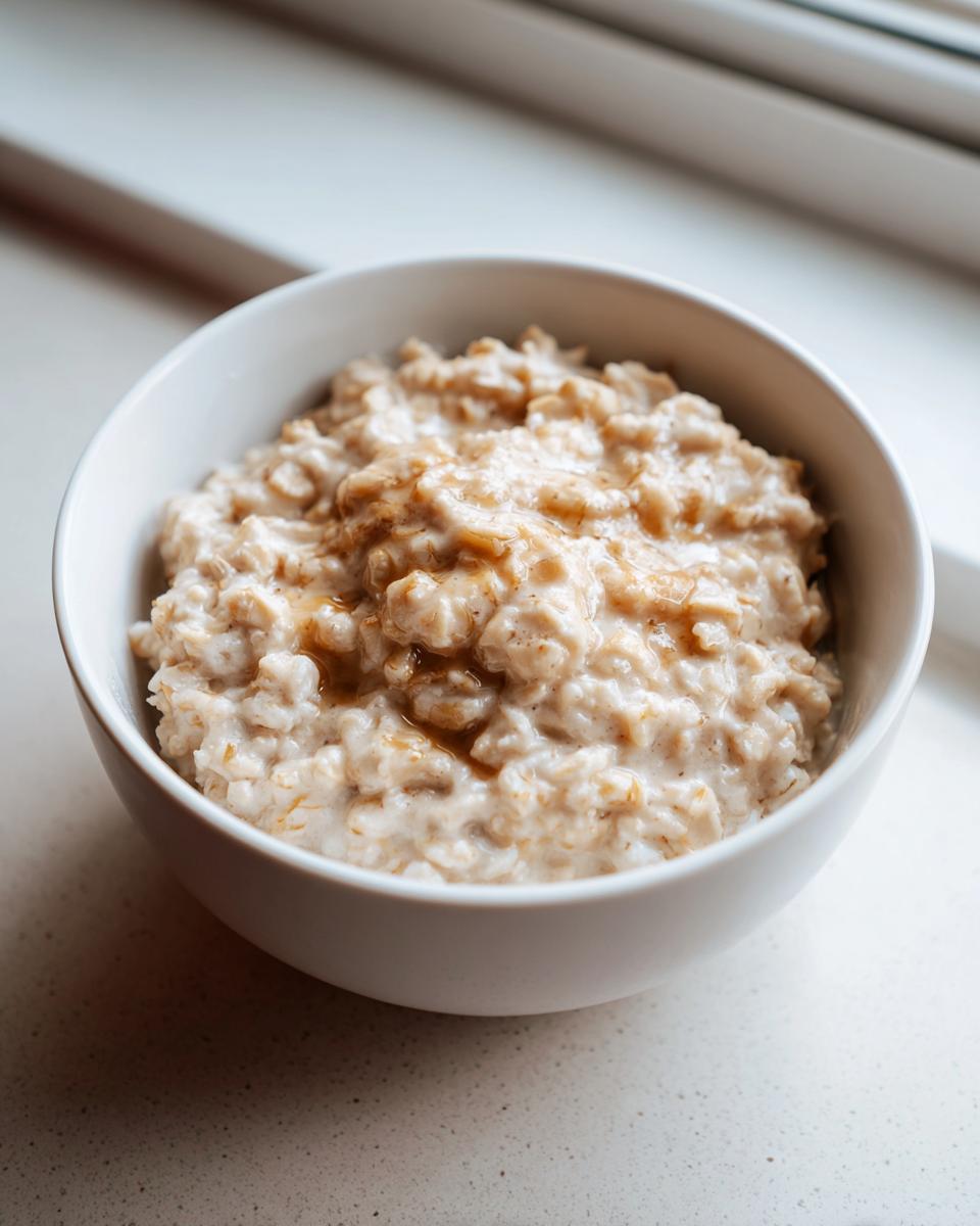Close-up of creamy banana oatmeal drizzled with syrup in a white bowl near a window.