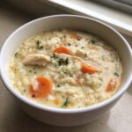 Close-up of a creamy chicken and carrot rice soup garnished with parsley in a white bowl.