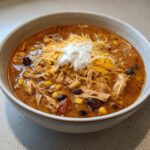A close-up of creamy chicken taco soup topped with shredded cheese and a dollop of sour cream.