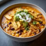 Close-up of a bowl of creamy chicken tortilla soup, loaded with shredded chicken, black beans, corn, and topped with avocado, sour cream, and tortilla strips.