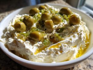 A white bowl filled with creamy dirty martini dip, topped with whole green olives, chopped peppers, and herbs.