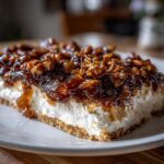 A thick slice of pecan pie dip served on a white plate, featuring a graham cracker crust, creamy layer, and sticky pecan topping.