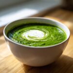 A close-up of vibrant green spinach soup topped with a swirl of cream and cracked black pepper.