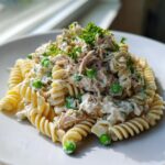 A serving of creamy tuna pasta salad made with rotini pasta, tuna, and bright green peas, topped with fresh parsley.
