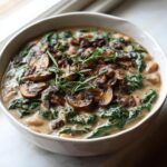 A close-up of a bowl of creamy white bean mushroom stew topped with sautéed mushrooms, spinach, and fresh rosemary.