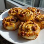 Five freshly baked sausage pinwheels topped with melted orange cheddar cheese on a white plate.