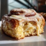 A close-up, mouth-watering shot of a single, fluffy cinnamon roll with a bite taken out, covered in thick white icing.
