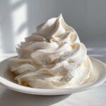 A tall, perfectly piped swirl of fluffy vanilla buttercream frosting in a small white bowl.
