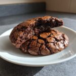 Two fudgy brownie cookies on a white plate, one broken in half revealing a molten center.