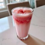A tall glass filled with frothy, pink strawberry milk featuring visible layers of crushed strawberries.