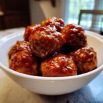 Close-up of several saucy, glazed cocktail meatballs piled high in a small white serving bowl.