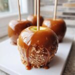 A close-up of a glossy caramel apple dipped in crushed nuts, with caramel dripping down the side.