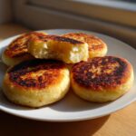 A stack of freshly cooked, golden brown irish potato farls on a white plate, one is broken open to show the texture.