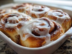 Close-up of freshly baked, soft cinnamon buns covered in thick white icing in a white baking dish.