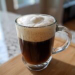 Close-up of a glass mug filled with dark Irish Coffee topped with a thick layer of light cream foam.