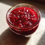Close-up overhead view of a glass jar filled with vibrant, rich red christmas jam, showing visible fruit seeds.