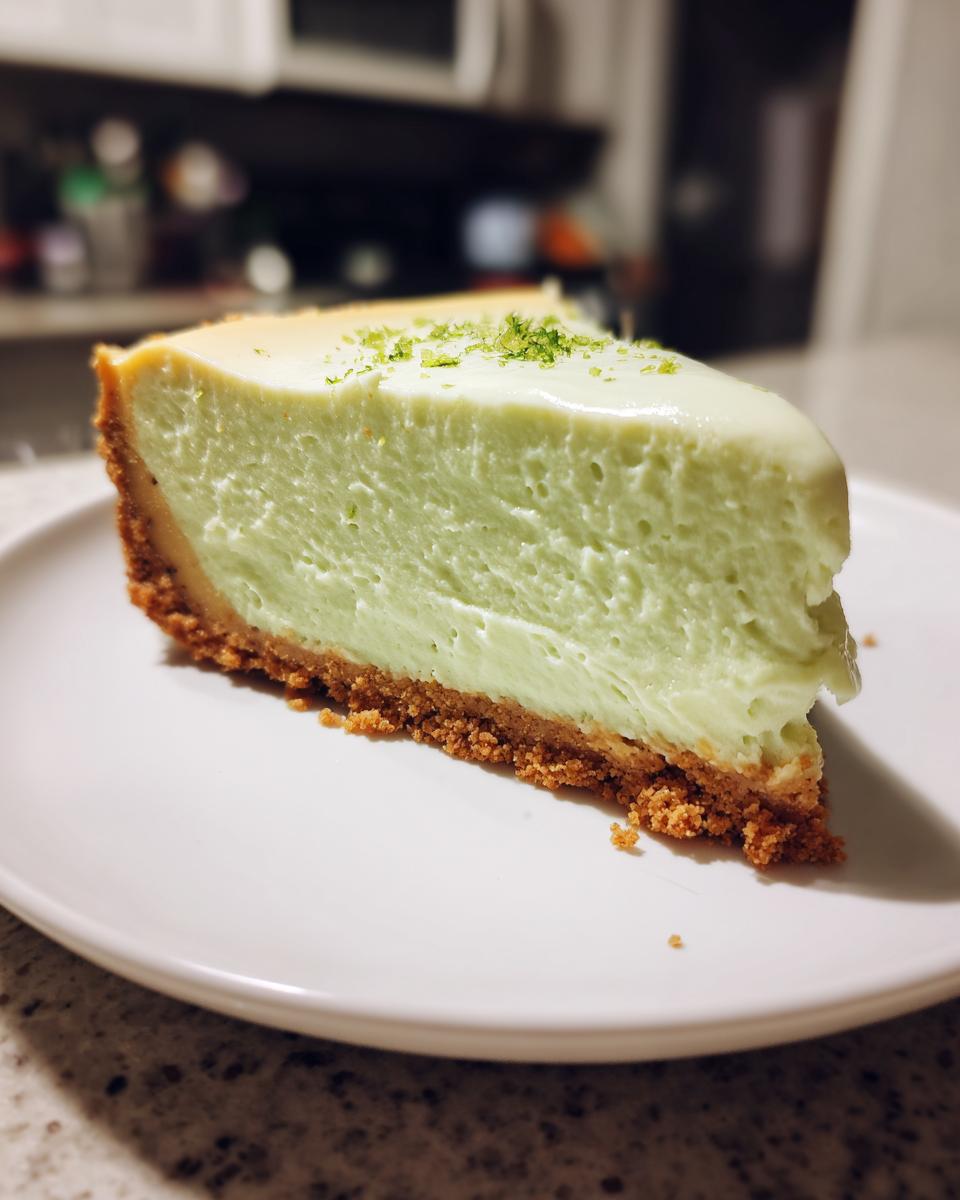 A close-up of a slice of light green key lime cheesecake on a white plate, topped with lime zest.