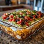 Close-up of a fully assembled Seven layer dip in a clear glass baking dish, topped with tomatoes, olives, and green onions.