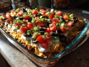 A vibrant, layered Seven layer dip topped with shredded cheese, sour cream, tomatoes, olives, and green onions.