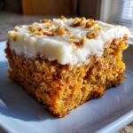 A close-up of a moist slice of carrot cake bars topped with thick cream cheese frosting and chopped walnuts.