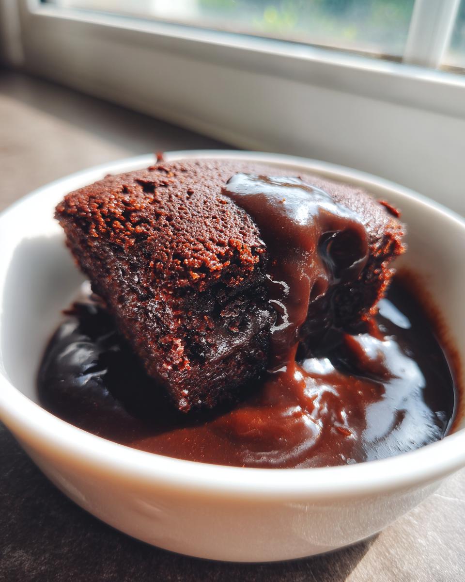 A close-up of a rich, dark chocolate pudding cake served in a white bowl, covered in warm chocolate sauce.