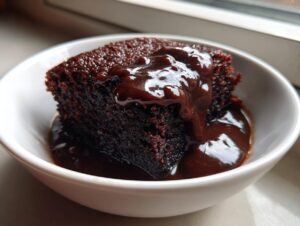 A rich slice of chocolate pudding cake topped with warm, gooey chocolate sauce served in a white bowl.