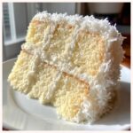 Close-up of a moist slice of coconut cake, layered with white frosting and covered in shredded coconut flakes.