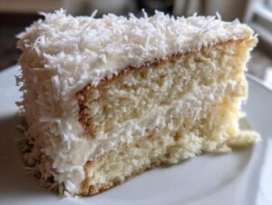 Close-up of a moist slice of coconut cake layered with white frosting and covered in shredded coconut.