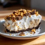 A generous slice of no-bake butterfinger pie featuring a dark crust, creamy filling, whipped topping, and crushed Butterfinger candy.