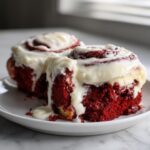 Two decadent red velvet cinnamon rolls heavily topped with dripping white cream cheese frosting on a white plate.