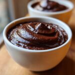 Close-up of thick, glossy chocolate pudding served in a white bowl, with another bowl blurred in the background.