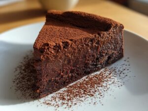 A decadent slice of flourless chocolate torte, intensely dark and moist, dusted heavily with cocoa powder on a white plate.