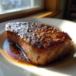 A thick, perfectly seared tuna steak resting in its dark marinade, topped with sesame seeds.