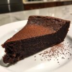 A decadent slice of flourless chocolate torte dusted heavily with cocoa powder on a white plate.