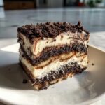 A layered slice of no-bake dirt cake featuring dark cookie crumbs, white cream, and ladyfingers on a white plate.
