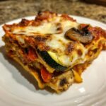 Close-up of a layered slice of roasted vegetable lasagna showing zucchini, mushrooms, and bubbly melted cheese.